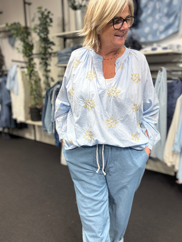 Smuk plus size bluse fra Sigensgaard med broderede guld blomster. Den er i 100% bomuld, som gerne må se lidt krøllet ud. Den har v-hals, lange ærmer som kan smøges op grundet knapåbning ved ærmets afslutning. Her ses den i en flot lys blå farve.