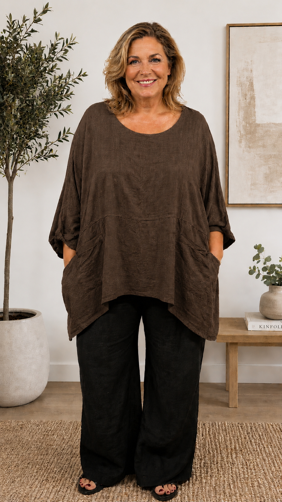 Kvinde i størrelse 46/48 iført Adler tunika som findes i flere farver. Tunikaen har en løs, oversized pasform med rund halsudskæring, 3/4-lange ærmer og store påsatte lommer foran. Den lette, strukturvævede kvalitet falder blødt over kroppen og giver et afslappet, komfortabelt look. Stylet efter din smag. Billed er Ai generet.