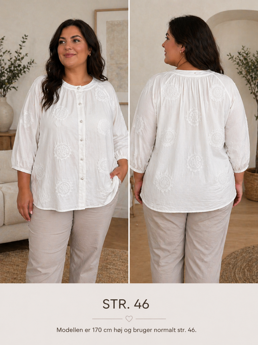Astrid skjorte i let, hvid kvalitet med fine broderede detaljer og dekorative perleknapper foran. Blusen har rund hals, let rynk ved skuldrene og en løs, komfortabel pasform med 3/4-ærmer.