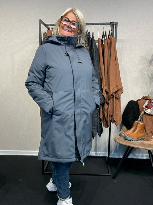 Smart og funktionel plus size jakke efterårs/vinter jakke hvor der er mulighed for at knappe en smule op i siden. Den er med lange ærmer, hætte og så har den lynlås lukning hele vejen ned, samt lommer med lynlås. Rigtig god længde som går til lige under knæet.