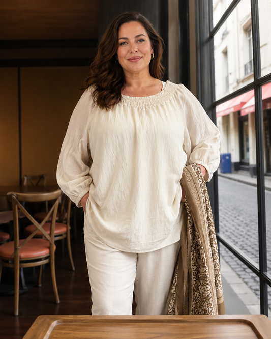 Molly bluse i sandfarvet nuance vist på en plus size-model i størrelse 46/48. Blusen har en feminin elastisk udskæring, lange ballonærmer med elastikkant og en løs, komfortabel pasform. Den lette kvalitet falder flot over kroppen og er stylet med lyse bukser. Billedet er AI-genereret.