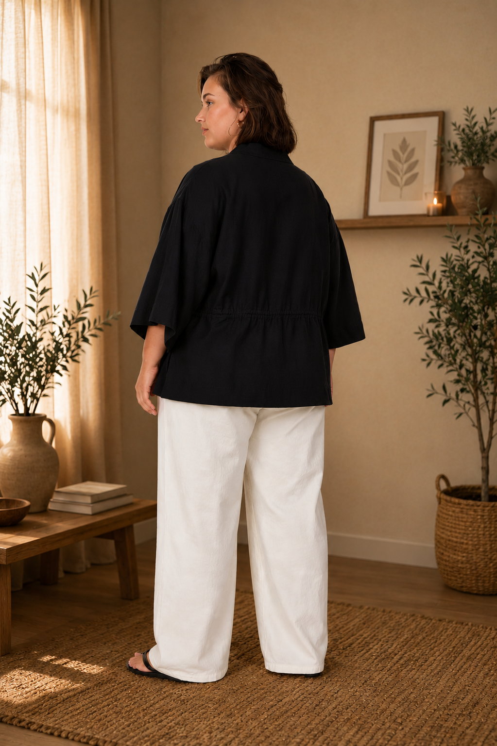 Kurvet model i størrelse 46 iført Corinna 25 kimono i sort. Kimonoen har en løs og afslappet pasform med brede ¾-ærmer og justerbar taljesnøre bagpå. Modellen er fotograferet bagfra i et lyst og hyggeligt interiør med varme naturtoner, træmøbler og grønne planter. Stylet med hvide, vide bukser og sandaler.