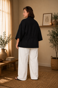 Kurvet model i størrelse 46 iført Corinna 25 kimono i sort. Kimonoen har en løs og afslappet pasform med brede ¾-ærmer og justerbar taljesnøre bagpå. Modellen er fotograferet bagfra i et lyst og hyggeligt interiør med varme naturtoner, træmøbler og grønne planter. Stylet med hvide, vide bukser og sandaler.