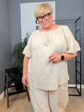 Ella tunika i hvid på plus size-model i størrelse 46/48. Tunikaen er fremstillet i en skøn, blød og let kvalitet med løs pasform, rund hals med bindebånd og dekorative kvaster samt feminine flæseærmer. Den luftige model giver et afslappet og komfortabelt udtryk og er stylet med jeans. Billedet er AI-genereret.