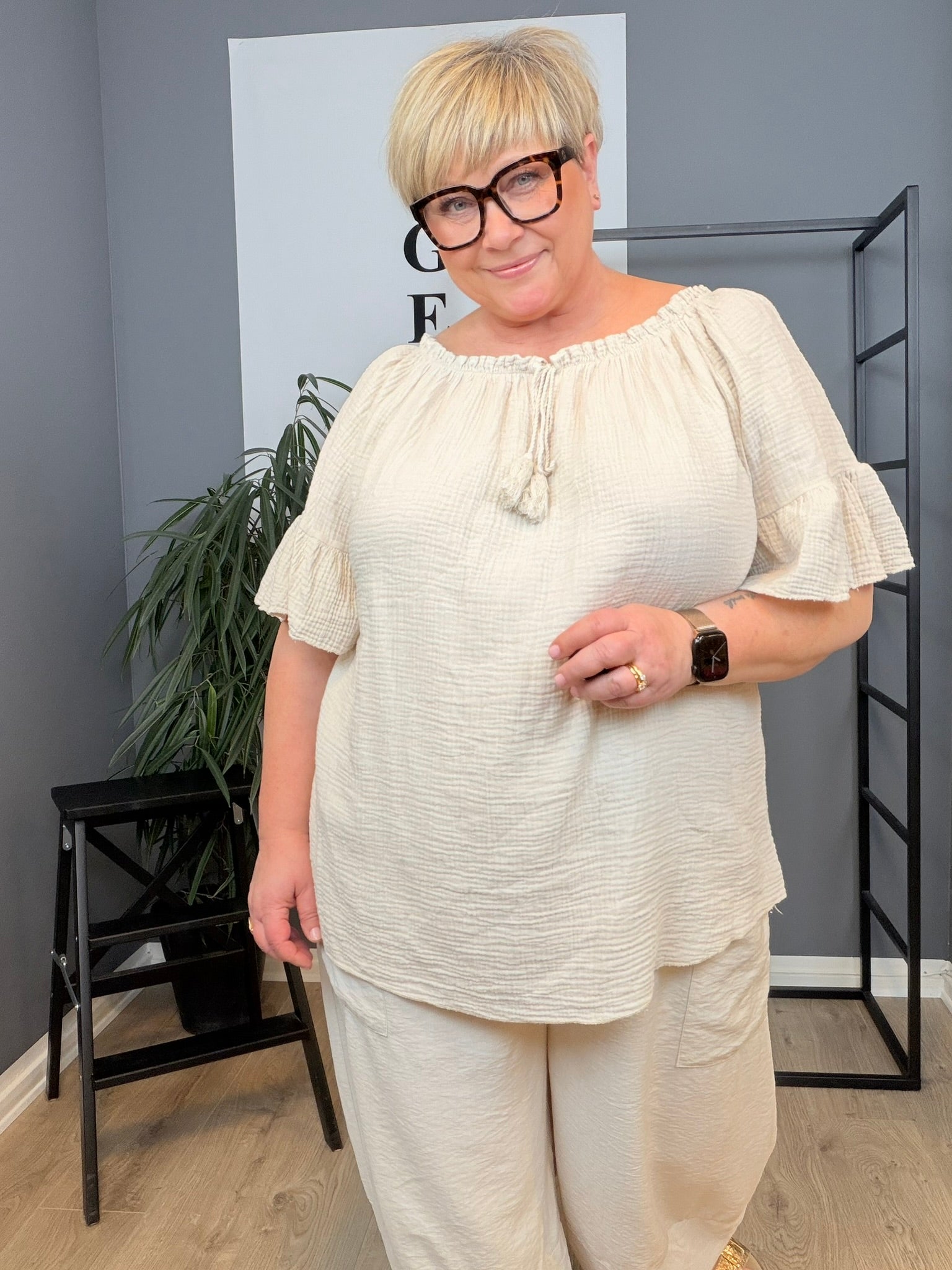 Ella tunika i hvid på plus size-model i størrelse 46/48. Tunikaen er fremstillet i en skøn, blød og let kvalitet med løs pasform, rund hals med bindebånd og dekorative kvaster samt feminine flæseærmer. Den luftige model giver et afslappet og komfortabelt udtryk og er stylet med jeans. Billedet er AI-genereret.