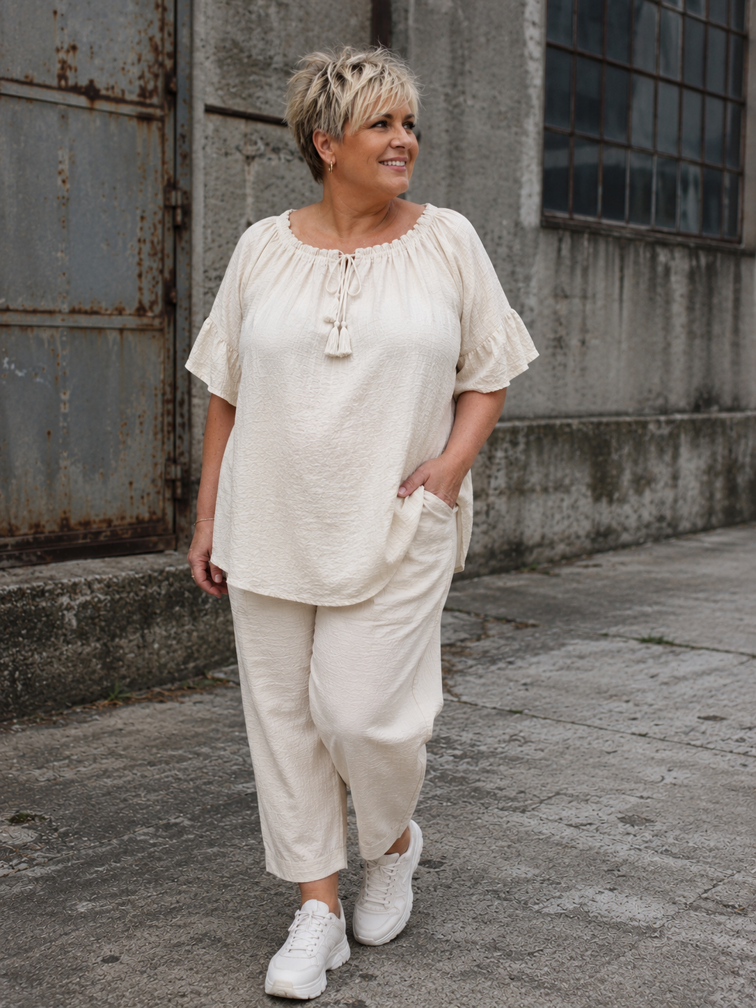 Ella tunika i hvid på plus size-model i størrelse 46/48. Tunikaen er fremstillet i en skøn, blød og let kvalitet med løs pasform, rund hals med bindebånd og dekorative kvaster samt feminine flæseærmer. Den luftige model giver et afslappet og komfortabelt udtryk og er stylet med jeans. Billedet er AI-genereret.