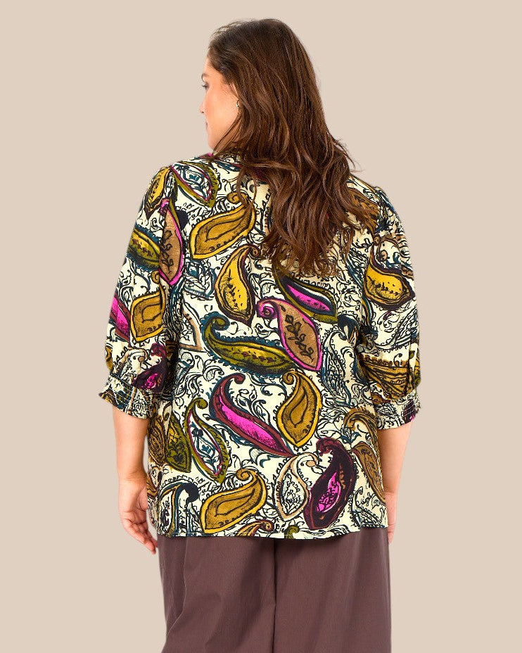Heva 2 viskose bluse fra Wasabi vist på en AI-genereret model i størrelse 46/48. Blusen har et dekorativt paisleymønster i beige, brun, olivengrøn og pink nuancer, V-hals og 3/4-lange ærmer med let vidde. Modellen er stylet med lyse bukser og står i et hyggeligt cafémiljø udendørs, som fremhæver blusens afslappede og feminine udtryk. Billedet er AI-genereret.