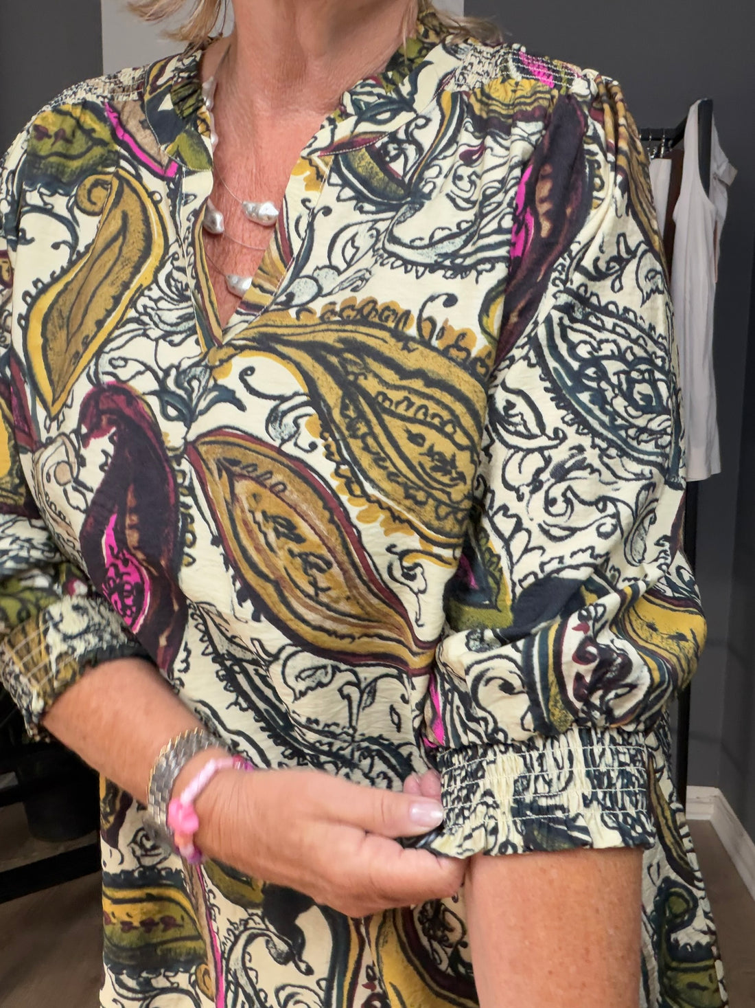 Heva 2 viskose bluse fra Wasabi vist på en AI-genereret model i størrelse 46/48. Blusen har et dekorativt paisleymønster i beige, brun, olivengrøn og pink nuancer, V-hals og 3/4-lange ærmer med let vidde. Modellen er stylet med lyse bukser og står i et hyggeligt cafémiljø udendørs, som fremhæver blusens afslappede og feminine udtryk. Billedet er AI-genereret.