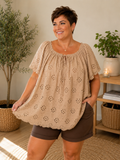 Janne blondebluse i taupe med smukke broderie anglaise-detaljer. Feminin plus size bluse med løs A-facon, elastisk halsudskæring med bindebånd og korte blondeærmer. Den lette bomuldskvalitet giver et luftigt og komfortabelt look, perfekt til både shorts, jeans og hørbukser på varme sommerdage. smart plussize overdel,billed er  ai generet.