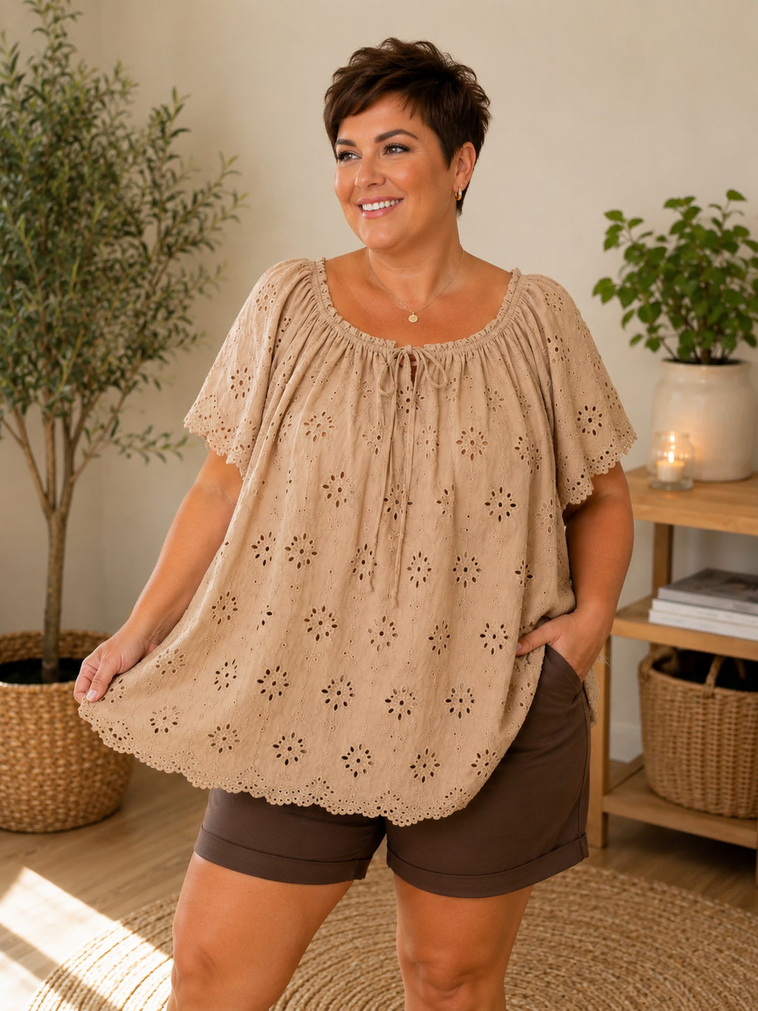 Janne blondebluse i taupe med smukke broderie anglaise-detaljer. Feminin plus size bluse med løs A-facon, elastisk halsudskæring med bindebånd og korte blondeærmer. Den lette bomuldskvalitet giver et luftigt og komfortabelt look, perfekt til både shorts, jeans og hørbukser på varme sommerdage. smart plussize overdel,billed er  ai generet.