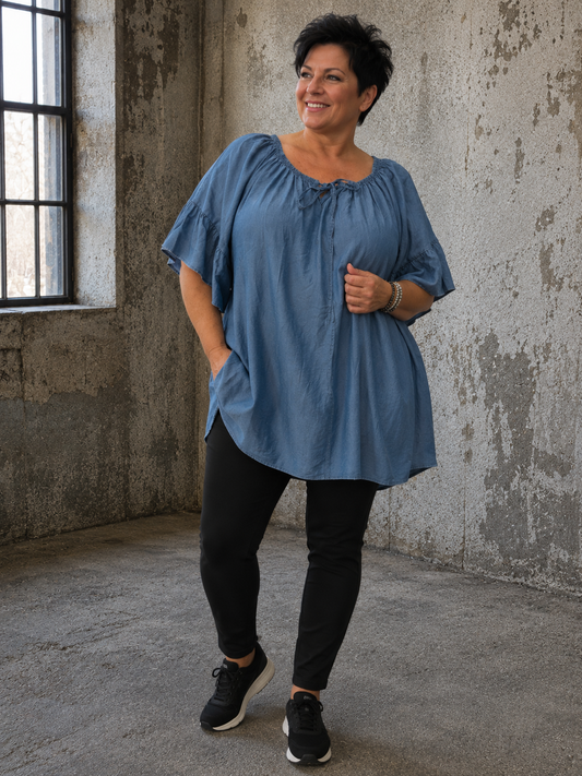 Joan er en skøn plussize tunika i blød tencel med et flot fald og en feminin pasform. Den brede elastiske halsudskæring med bindebånd og de fine flæseærmer giver tunikaen et elegant og afslappet udtryk, som passer perfekt til både hverdag og fest. billed er ai generet.