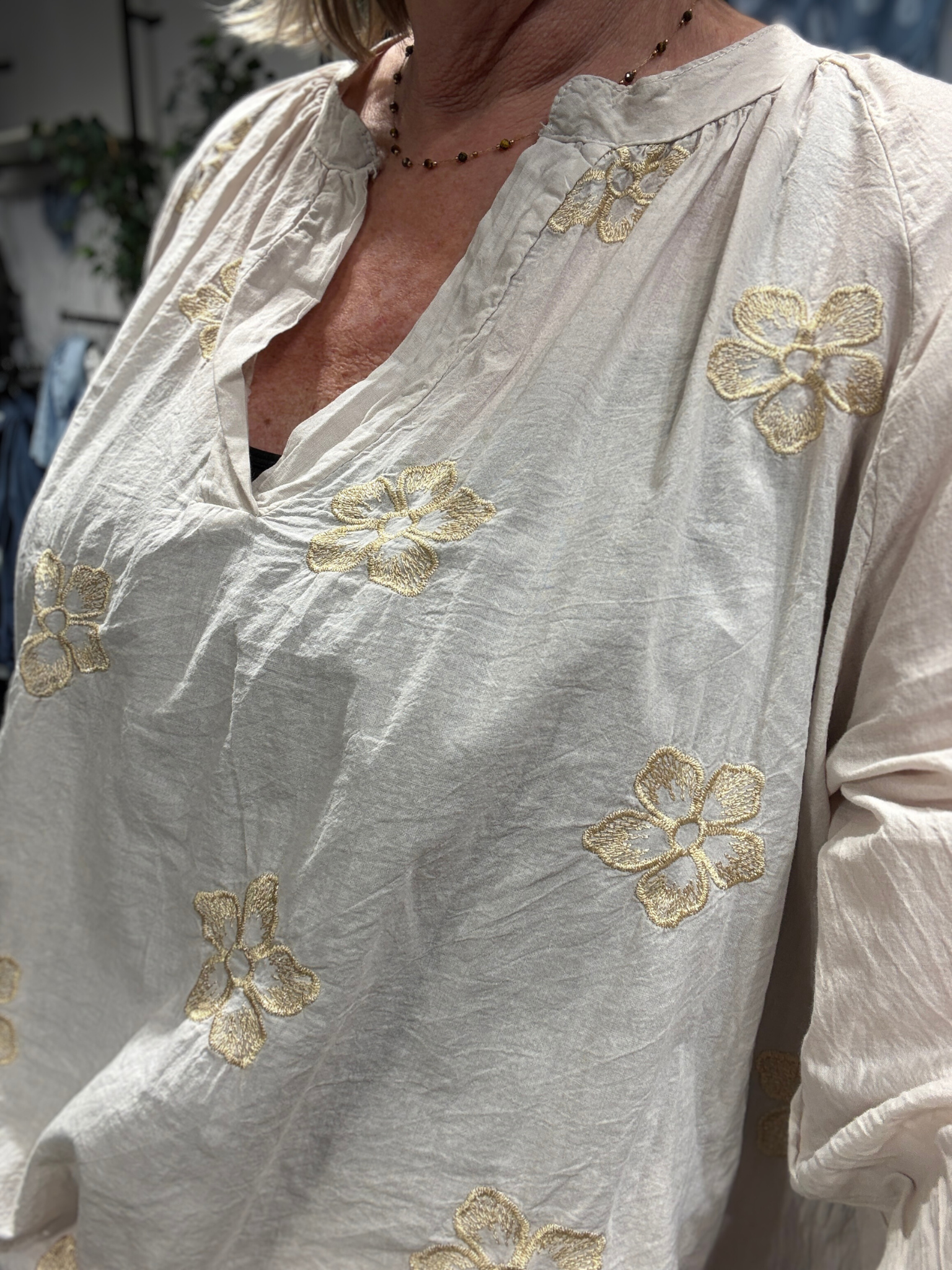 Smuk plus size bluse fra Sigensgaard med broderede guld blomster. Den er i 100% bomuld, som gerne må se lidt krøllet ud. Den har v-hals, lange ærmer som kan smøges op grundet knapåbning ved ærmets afslutning. Her ses den i en flot beige farve.