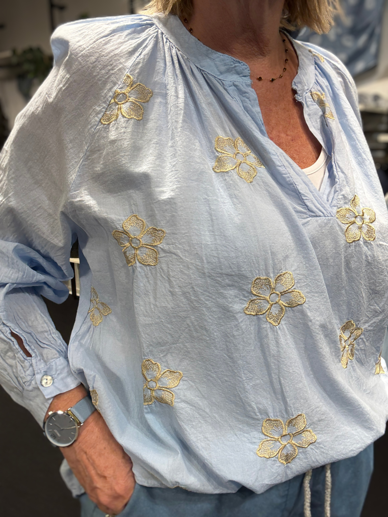 Smuk plus size bluse fra Sigensgaard med broderede guld blomster. Den er i 100% bomuld, som gerne må se lidt krøllet ud. Den har v-hals, lange ærmer som kan smøges op grundet knapåbning ved ærmets afslutning. Her ses den i en flot lys blå farve.