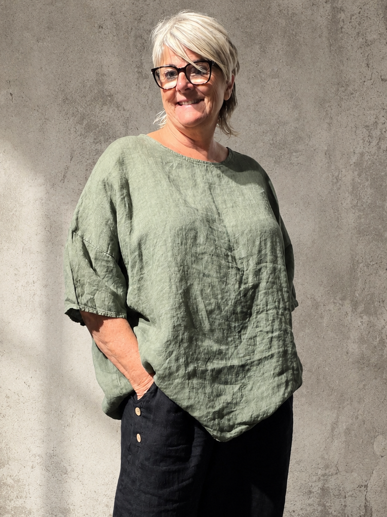 Linda hørtunika til pluspiger – rummelig tunika i hør med afslappet pasform, stylet med sorte bukser i et råt og naturligt butiksmiljø.