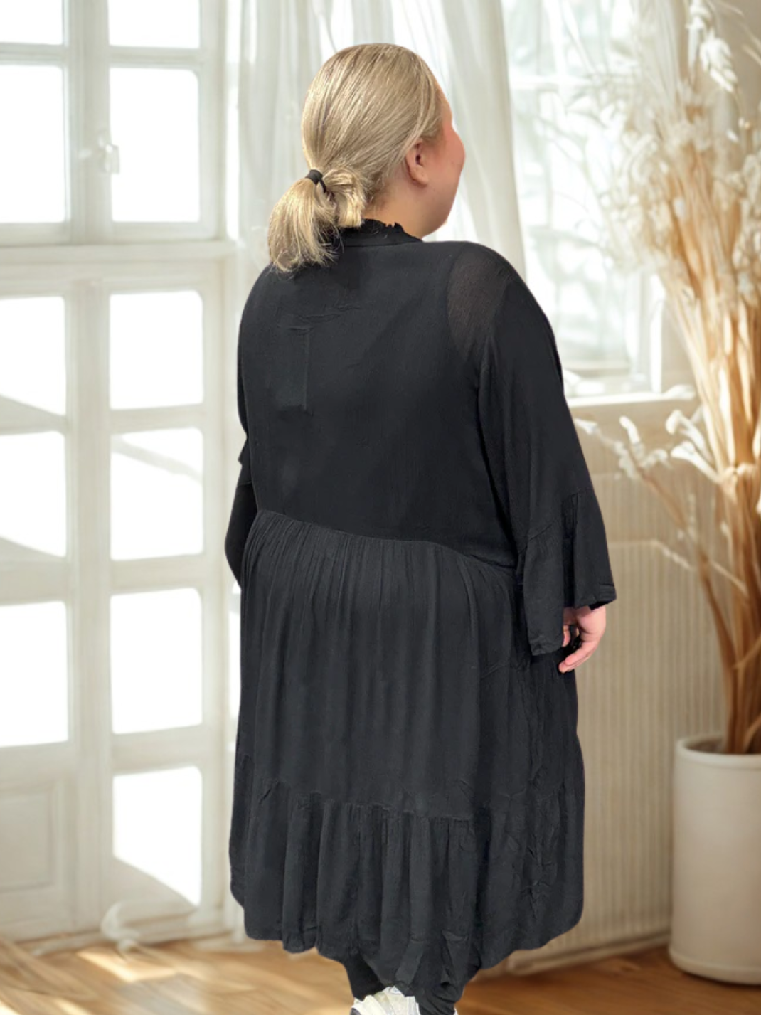 Pyntet sort kjole som er i en god facon til plussize kvinder. Den er fra mærket Kaffe Curve, og modellen på billedet bruger en str. 54. 

*Baggrunden på billedet er AI-genereret