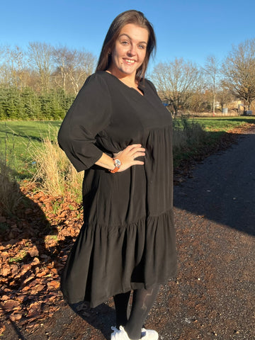 "Rigmor plussize-kjole med flot A-facon, flere lette lag, forlænget ryg og to diskret placerede lommer foran, der virker slankende."