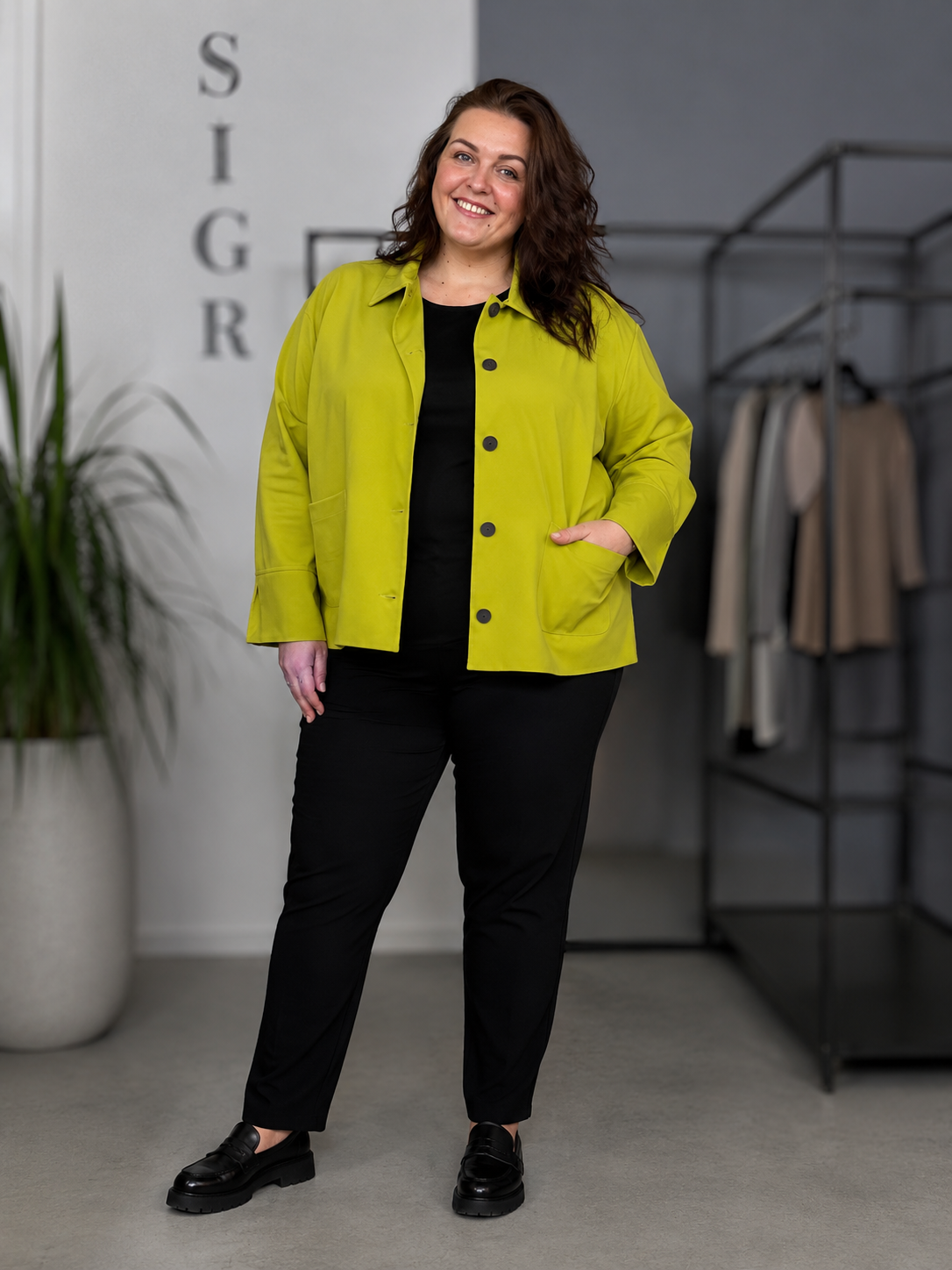 Kvinde i plus size (str. 46/48) iført limegrøn Sally 1 jakke fra Wasabi over sort bluse og sorte bukser. Modellen er 168 cm høj og står i et lyst showroom med et afslappet smil og hånden i lommen. Jakken har klassisk krave, mørke knapper og store påsatte lommer. billed er ai generet