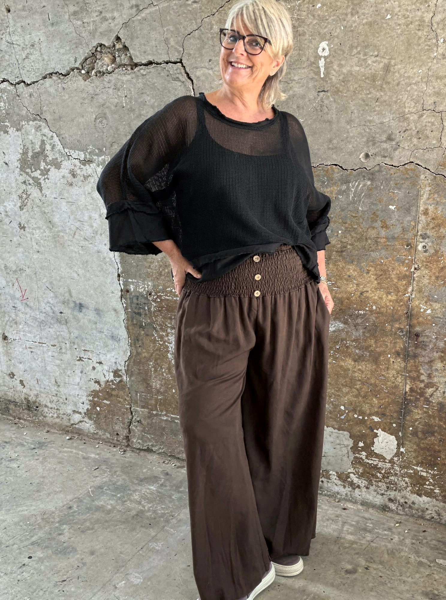 "Model i Tora viddebuks med bred smock-elastik i taljen, dekorative knapper og vide ben. Buksen vises i brun  sammen med en løs camel bluse. Modellen er str. 46/48 og 168 cm høj." billed er ai generet