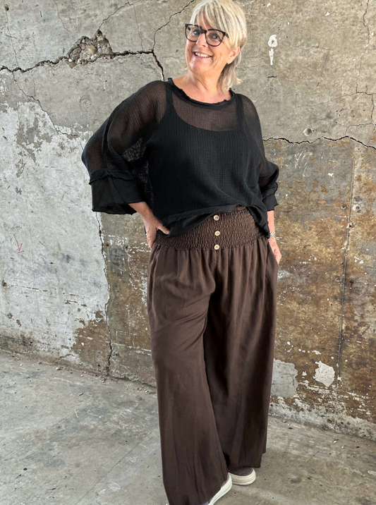 "Model i Tora viddebuks med bred smock-elastik i taljen, dekorative knapper og vide ben. Buksen vises i brun  sammen med en løs camel bluse. Modellen er str. 46/48 og 168 cm høj." billed er ai generet