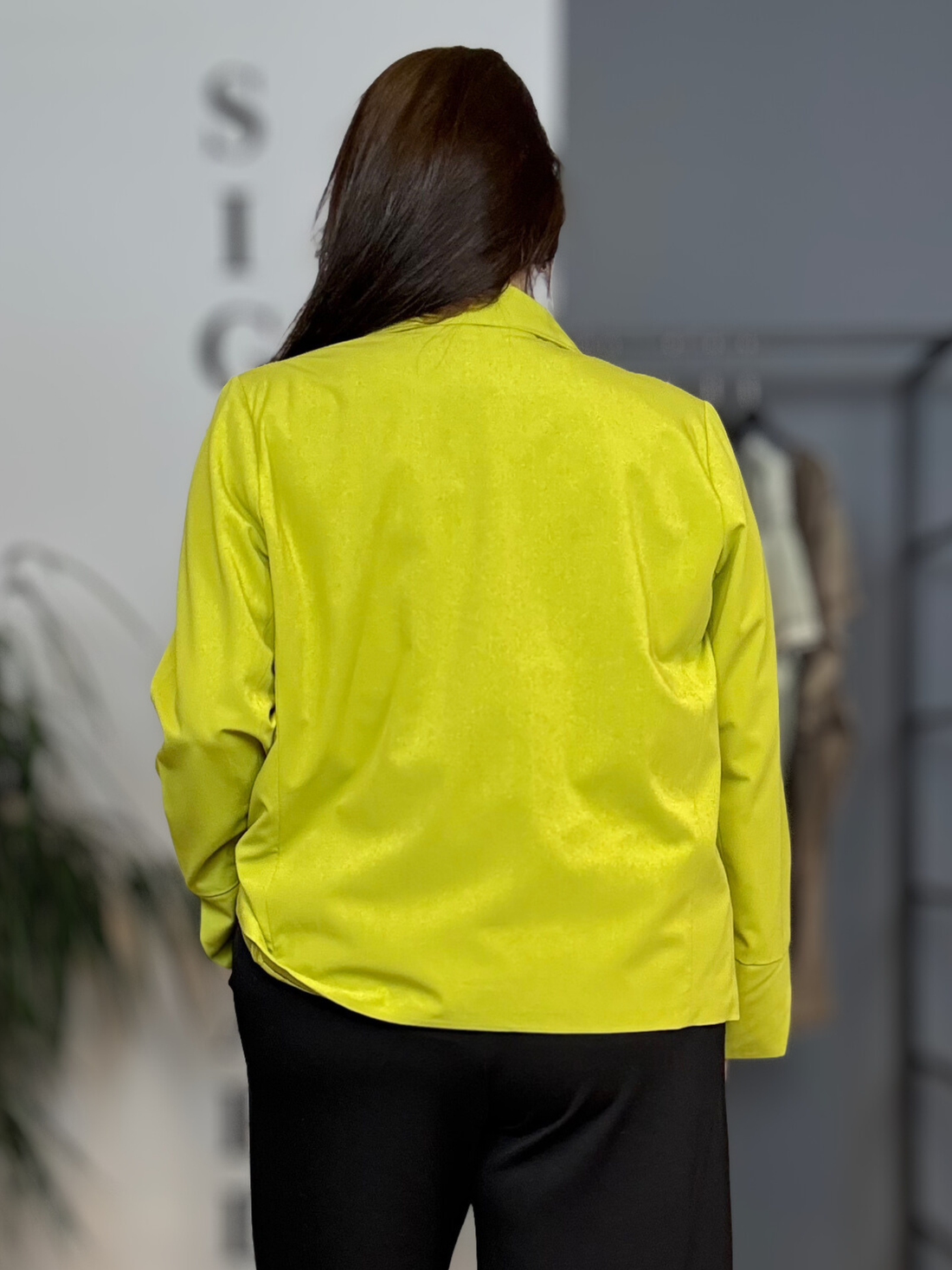 Kvinde i plus size (str. 46/48) iført limegrøn Sally 1 jakke fra Wasabi over sort bluse og sorte bukser. Modellen er 168 cm høj og står i et lyst showroom med et afslappet smil og hånden i lommen. Jakken har klassisk krave, mørke knapper og store påsatte lommer. billed er ai generet