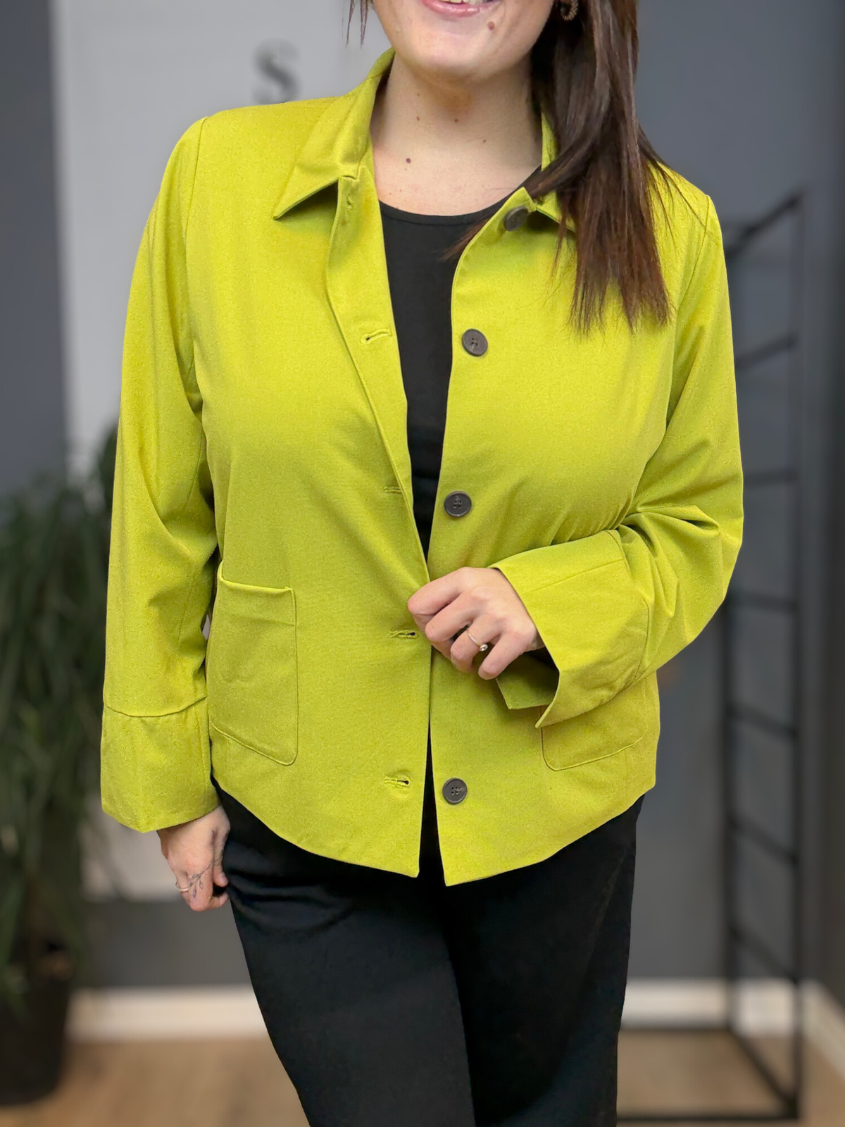 Kvinde i plus size (str. 46/48) iført limegrøn Sally 1 jakke fra Wasabi over sort bluse og sorte bukser. Modellen er 168 cm høj og står i et lyst showroom med et afslappet smil og hånden i lommen. Jakken har klassisk krave, mørke knapper og store påsatte lommer. billed er ai generet