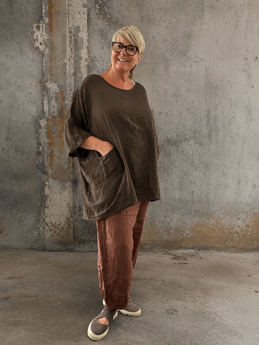 **Alt-tekst – Susanne plussize bomuldstunika (AI-genereret billede):**

AI-genereret billede af en smilende plus-size model iført Susanne bomuldstunika i mørk brun. Tunikaen har en løs, oversize pasform med rund hals, korte brede ærmer og store påsyede lommer foran. Den er stylet med rustfarvede bukser og komfortable sko i et råt industrielt miljø, som fremhæver tunikaens afslappede og moderne udtryk. Ideel til kvinder, der ønsker komfort, bevægelsesfrihed og et stilfuldt hverdagslook.
