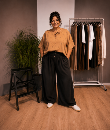 "Model i Tora viddebuks med bred smock-elastik i taljen, dekorative knapper og vide ben. Buksen vises i sort sammen med en løs brun bluse. Modellen er str. 46/48 og 168 cm høj." billed er ai generet