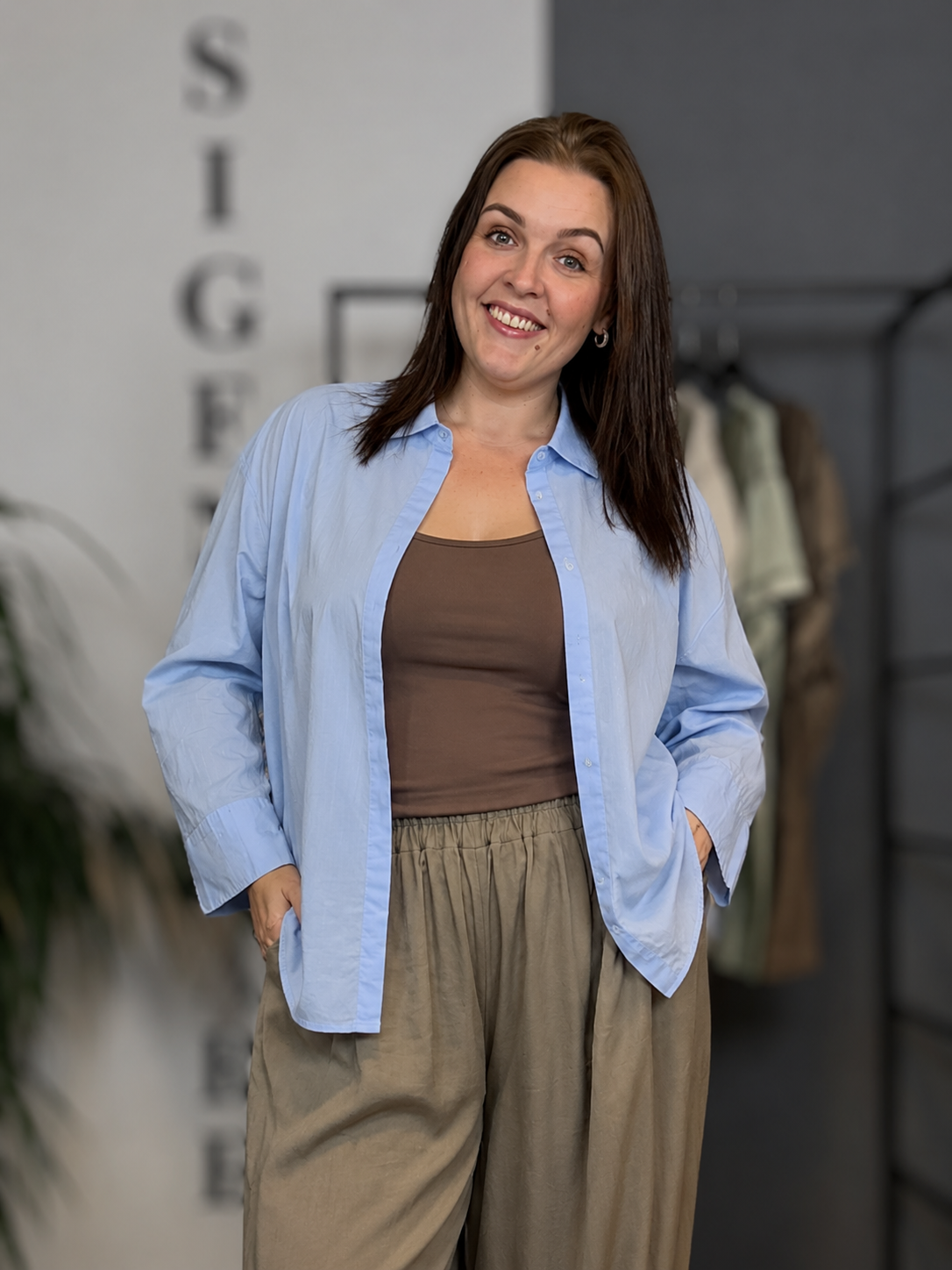 Mary 8 Plus Size skjorte i lyseblå med klassisk krave og lange ærmer. Skjorten bæres åben over en brun top og har en løs, komfortabel pasform. Vist på model i størrelse 46/48.